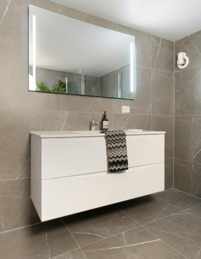 Ein modernes Badezimmer mit dunklen Großformatfliesen in Natursteinoptik, hängendem Waschtisch und großem, beleuchtetem Spiegel.