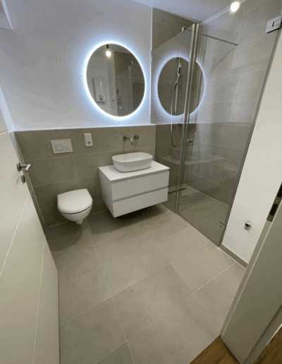 Ein modernes Badezimmer mit naturfarbenen Fliesen, beleuchtetem Spiegel, hängendem Waschbecken mit wandmontierter Armatur, Hänge-WC und Glasdusche.